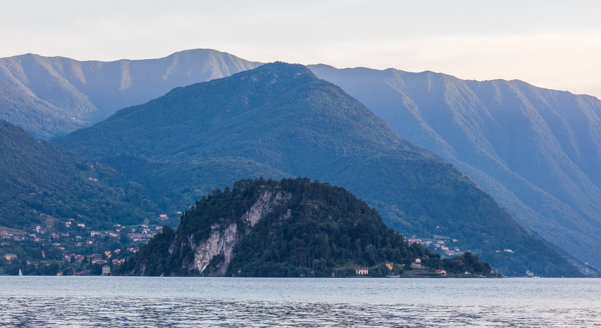 Blue mountains rising above a calm lake, with a small lakeside village and a wooded headland in the foreground.