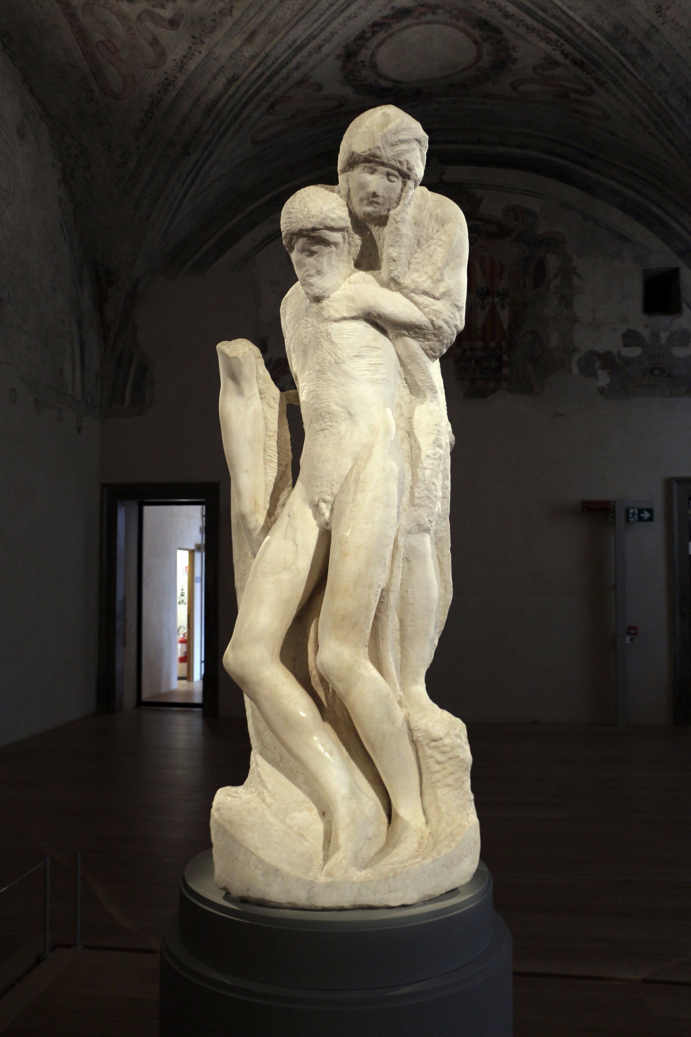 Tall white marble sculpture of two intertwined figures on a dark plinth in a stone-walled room.