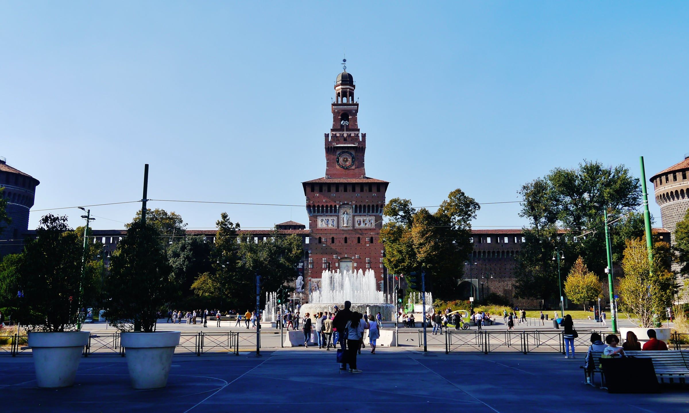Brick front of Sforza Castle with its tall Filarete Tower rising above a broad square, fountains and trees in front.